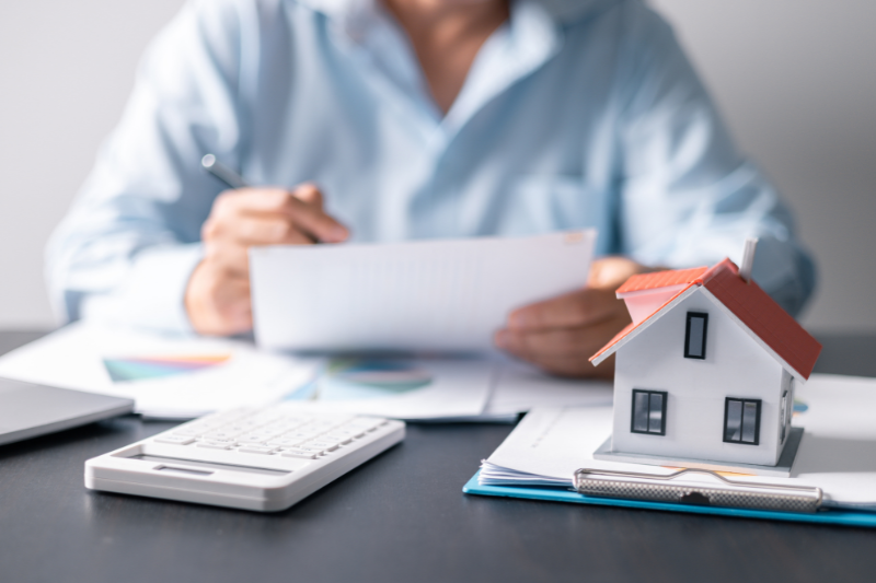 A person sits at a desk reviewing documents with charts, next to a calculator and a model house, suggesting work related to real estate or financial planning.