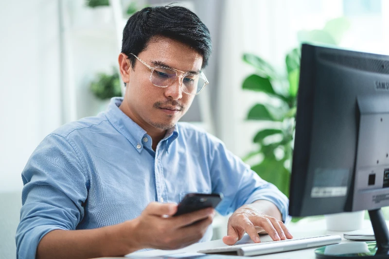 A man wearing glasses is sitting at a desk with a computer and a cell phone